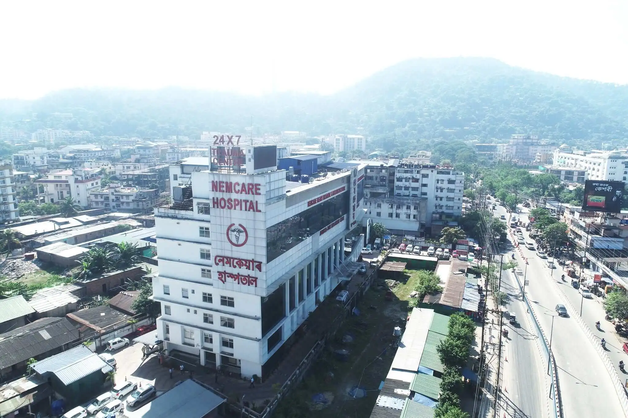 Aerial view of a NEMCARE Hospital building in Assam, with its name in English and Assamese on the front and a 24x7 trauma centre sign on the roof